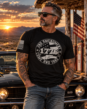 Man wearing FreedomBuilt 2nd Amendment 1776 Come and Take It graphic tee in Black Heather, printed with soft water-based ink, standing beside a classic black Camaro at sunset.
