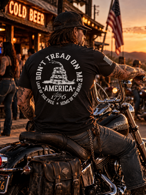 Rugged biker wearing a black FreedomBuilt Don't Tread on Me graphic tee with white rattlesnake and America 1776 back print, sitting on a Harley-Davidson motorcycle outside a biker bar at sunset.