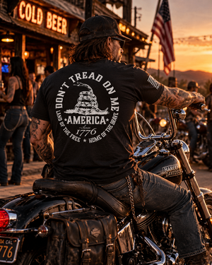 Rugged biker wearing a black FreedomBuilt Don't Tread on Me graphic tee with white rattlesnake and America 1776 back print, sitting on a Harley-Davidson motorcycle outside a biker bar at sunset.