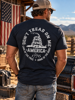 Man wearing a FreedomBuilt Don't Tread on Me navy blue graphic tee featuring a white rattlesnake and America 1776 back print, standing beside a vintage pickup truck.