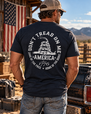 Man wearing a FreedomBuilt Don't Tread on Me navy blue graphic tee featuring a white rattlesnake and America 1776 back print, standing beside a vintage pickup truck.
