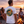 A man standing on a beach at sunset wearing a white t-shirt with a large circular 'Nautilus' graphic on the back, with two women sitting nearby by a campfire.