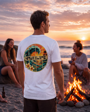 A man standing on a beach at sunset wearing a white t-shirt with a large circular 'Nautilus' graphic on the back, with two women sitting nearby by a campfire.